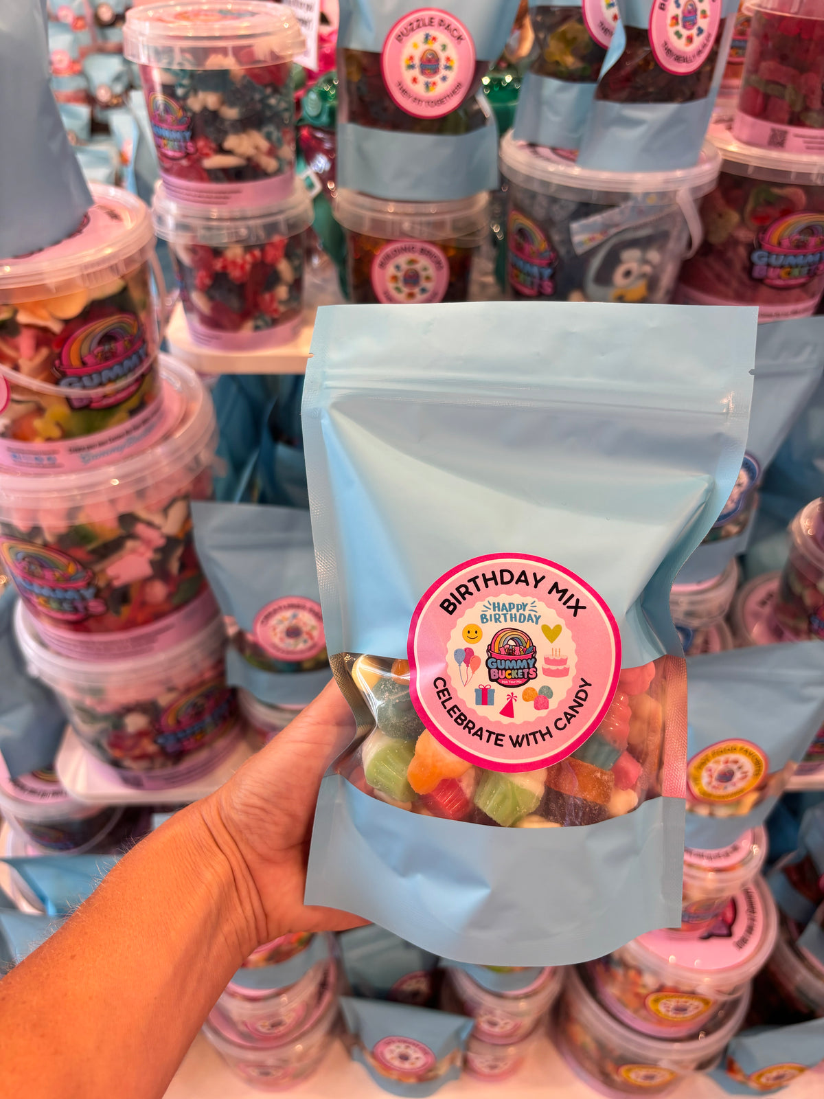 A hand holds a light blue stand-up pouch of 'Birthday Mix' gummy candy from Candy Cane Lane, with stacks of similar candy containers in the background.