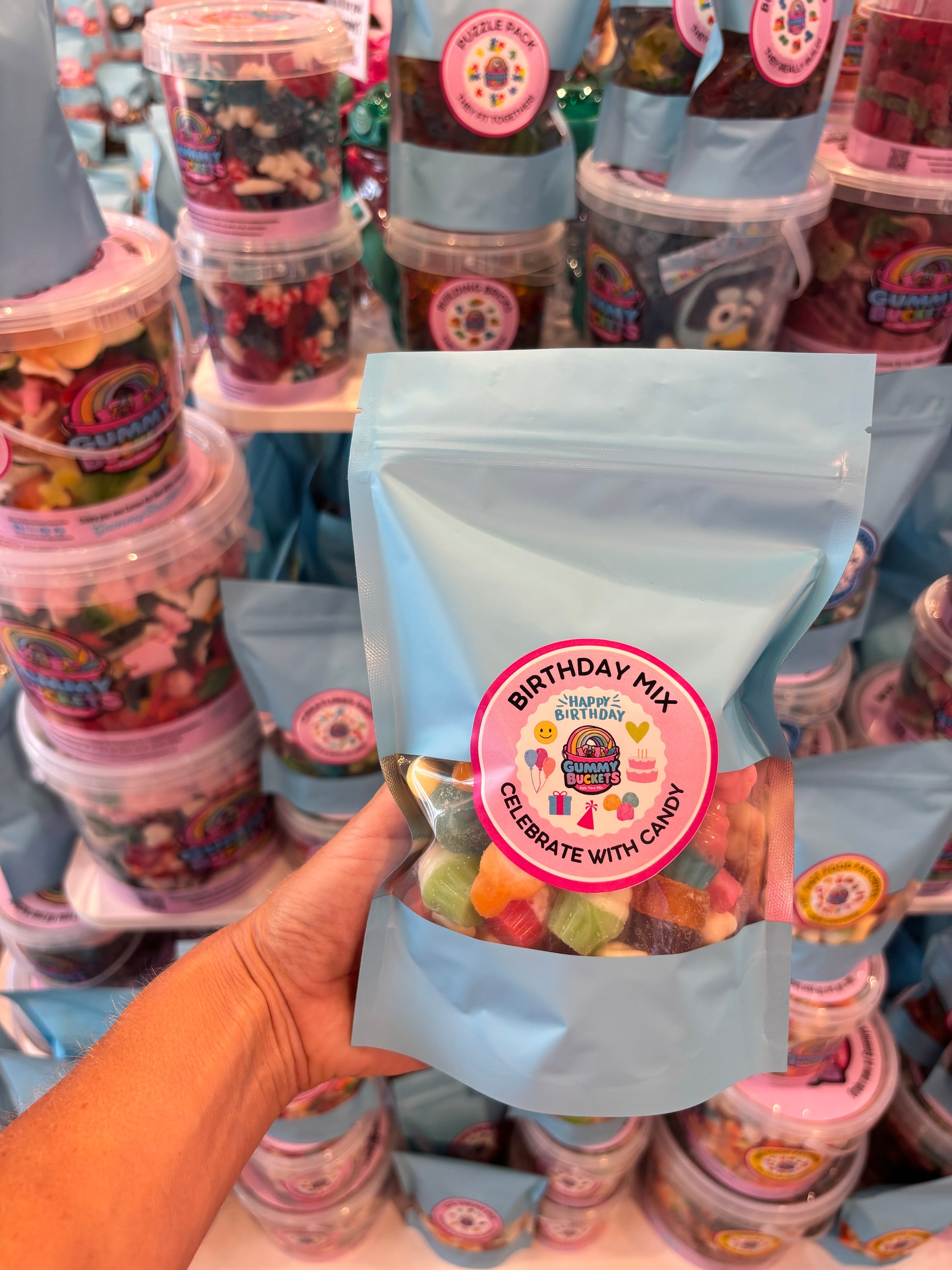 A hand holds a light blue stand-up pouch of 'Birthday Mix' gummy candy from Candy Cane Lane, with stacks of similar candy containers in the background.