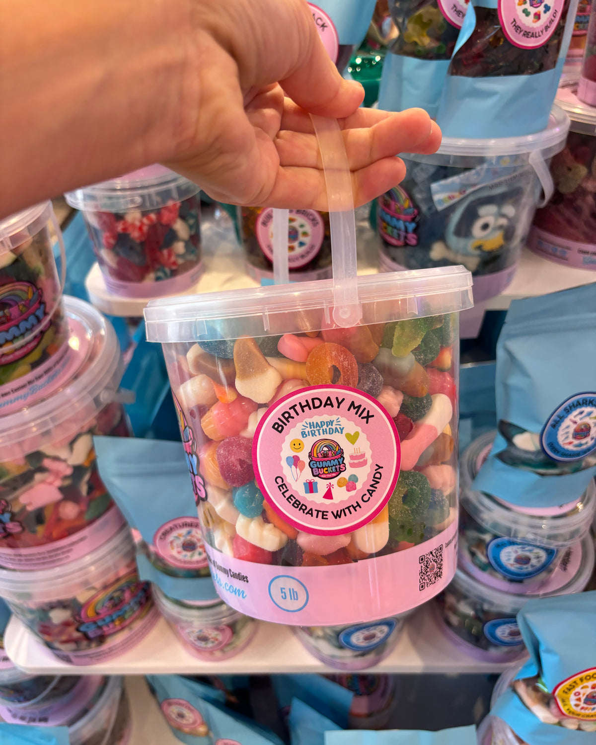 A hand holds a clear plastic tub filled with assorted colorful gummy candies and sweets. The tub has a pink label that reads 'BIRTHDAY MIX' and is shown on a store shelf surrounded by other candy containers.