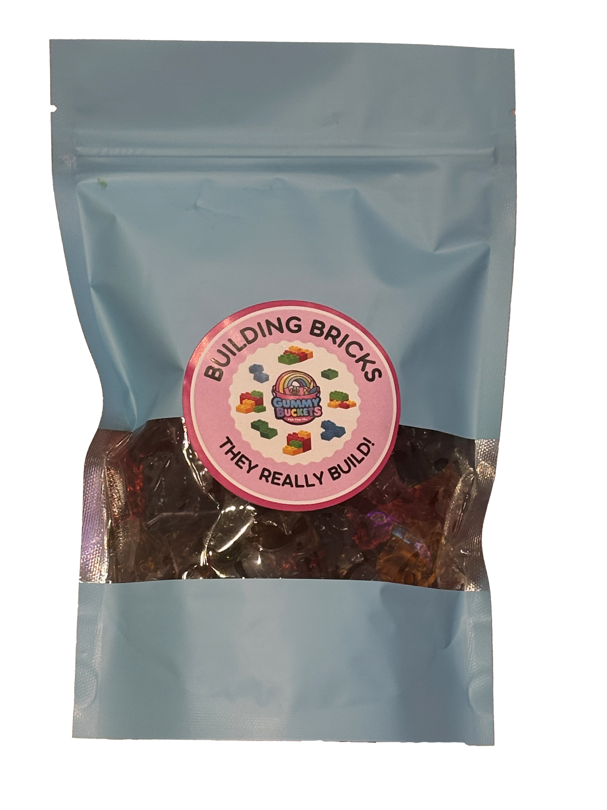 A light blue resealable stand-up pouch with a clear window showing colorful building brick-shaped gummy candies inside. A round pink label on the front reads 'BUILDING BRICKS' and 'THEY REALLY BUILD!' with the Gummy Buckets logo surrounded by illustrated gummy brick icons in the center.