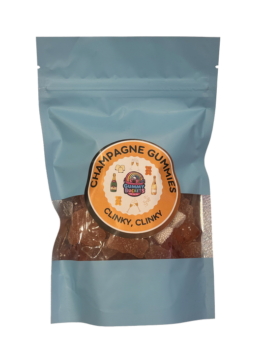 A light blue resealable stand-up pouch with a clear window showing golden amber champagne-flavored gummy bears inside. A round orange label on the front reads 'CHAMPAGNE GUMMIES' and 'CLINKY, CLINKY' with the Gummy Buckets logo surrounded by illustrated champagne bottles and glasses in the center.