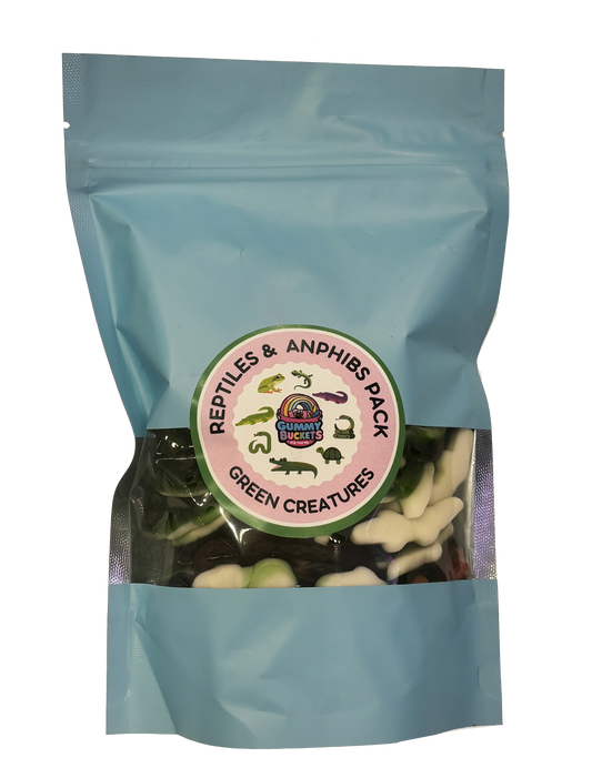 A light blue resealable stand-up pouch with a clear window showing green and white reptile and amphibian-shaped gummy candies including frogs, alligators, snakes, and turtles inside. A round green and pink label on the front reads 'REPTILES & AMPHIBS PACK' and 'GREEN CREATURES' with the Gummy Buckets logo surrounded by illustrated reptile silhouettes in the center.